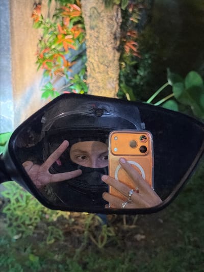 A person wearing a black helmet is reflected in a motorcycle's rearview mirror, taking a selfie with an orange smartphone. The person is making a peace sign with their left hand. They are wearing two rings on their right hand, one appears to be a diamond ring. The background is blurred, showing foliage and a textured tree trunk. The scene is dimly lit, suggesting it is nighttime. The image was taken in Kerambitan, Indonesia.