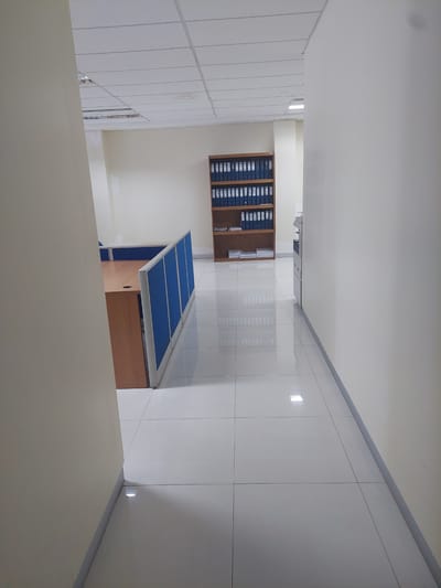 The image depicts a sterile, unoccupied office hallway with a tiled floor. To the left, a section of an office cubicle with a wooden desk and blue fabric dividers is visible. In the background, a tall wooden bookshelf is filled with numerous blue binders. Further down the hallway, a copier or printer is positioned against the right wall. The walls and ceiling are painted a neutral color, and fluorescent lights are embedded in the ceiling. The environment appears to be a modern office space in the City of Pematang Siantar, Indonesia, devoid of any activity or individuals.
