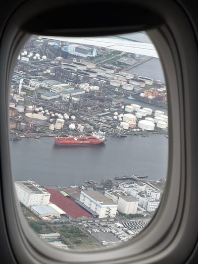 This aerial view captures an industrial area within the city of Kawasaki, Japan, as seen through an airplane window. The scene is dominated by a large harbor filled with industrial infrastructure. Prominently featured is a large, red tanker ship labeled "AURORA TANKERS," docked in the water.

Surrounding the harbor are numerous industrial facilities, including a complex array of storage tanks, some spherical and others cylindrical. These tanks are clustered together, indicating a significant industrial operation, likely related to oil or chemical storage and processing. Buildings of various sizes, including large warehouses and smaller administrative structures, are scattered throughout the industrial landscape. Some of these buildings have distinct color schemes, such as a white building with blue accents and another with a long red roof.

Beyond the immediate industrial zone, more buildings and what appear to be extensive parking lots filled with numerous vehicles, possibly trucks or containers, are visible. The overall environment suggests a heavily industrialized port city. The water in the harbor appears murky and dark. The image is framed by the distinct oval shape of an airplane window, with the wing of the aircraft visible at the top of the frame. The sky appears overcast.