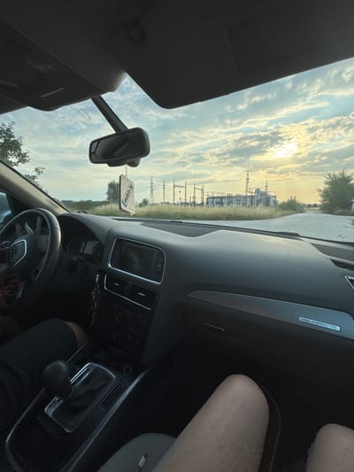 The image captures the interior of an Audi vehicle, specifically an SUV model, with a view through the windshield of an outdoor scene in Tsaratsovo, Bulgaria.
Inside the vehicle, a driver's hand is visible on the left side of the steering wheel, and their legs, clad in dark shorts or pants, are in the lower-left portion of the frame. An automatic transmission gear shifter is centrally located between the front seats. Keys are hanging near the center console. The dashboard features an infotainment screen, climate controls, and a "quattro" emblem on the passenger side trim. The legs of a passenger are visible in the lower-right foreground. A rearview mirror is suspended from the windshield, from which hangs a white air freshener displaying "mobile.bg" in green text. The vehicle's headliner and sun visors are visible across the top of the interior.
Outside the vehicle, the sky is partially covered with white, scattered clouds. The sun is low on the horizon, positioned towards the right side of the frame, indicating either sunrise or sunset and casting a soft, golden light. A large electrical power substation, comprising multiple metal structures, transformers, and power lines, is prominently visible in the distance. The ground leading to the substation appears unpaved, possibly a gravel or dirt road, and is flanked by overgrown grass and sparse foliage.