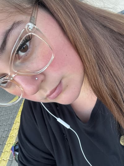 A woman with brown hair, wearing glasses and a black shirt, is taking a selfie outdoors. She has a nose piercing and is wearing earbuds. The background shows a yellow line on a paved surface, which could be a sidewalk or a street. The image does not contain information about the city of Bad Säckingen, Germany.