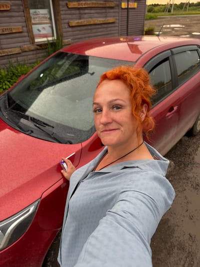 This media file shows a woman with vibrant red, curly hair posing for a selfie next to a red car. She is wearing a light blue, textured jacket and a dark necklace. Her expression is a slight smile with a playful tilt of her head. In her right hand, she holds a white cigarette with a purple and white filter pack. The car's hood is visible, showing a few water droplets, suggesting it might have recently rained or is a damp day.
In the background, a weathered wooden fence with vertical planks is visible. Attached to the fence is a rectangular sign with some text, and below it, another sign with Russian text, including the word "АНТЕННЫ" (ANTENNAS) and possibly other words related to sales or services. Further to the left, a narrow grassy area with trees can be seen. The overall lighting suggests it is daytime, possibly late afternoon given the warm tones. The scene appears to be outdoors, possibly in a rural or semi-rural setting in Sovkhoznyi, Russia, given the wooden structures and the language on the signs. The presence of the car and the woman's casual attire suggest a moment of personal time or a stop during a journey.