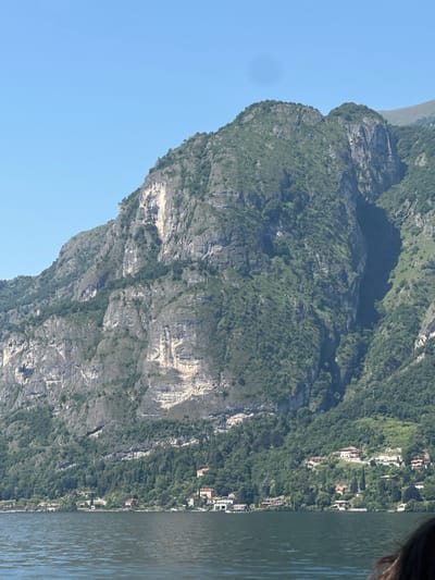 The image shows a wide shot of a steep, tree-covered mountain overlooking a body of water, likely Lake Como, with a few buildings nestled on the hillside in Varenna, Italy. The mountain is rugged and rocky in places, with a dense green forest covering most of its slopes. The water in the foreground is calm and dark, reflecting the sky and the mountains. The buildings appear to be villas or houses, with red-tiled roofs and white or light-colored walls. The sky is clear and blue.