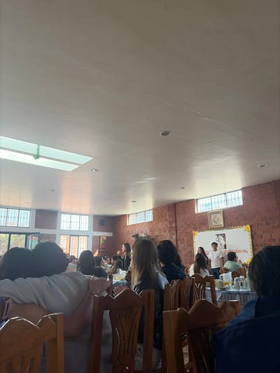 The scene is inside a building in Santa María Zacatepec, Mexico, where a group of people are seated in wooden chairs, possibly attending a lecture or presentation. Some individuals are interacting with mobile devices. At the front, there are speakers visible with a framed image or poster behind them. The building has brick walls, and multiple windows provide natural light.