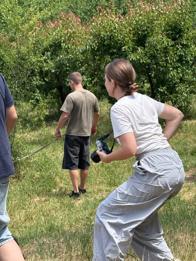 An outdoor scene in Grocka Urban Municipality, Serbia, depicts three individuals engaged in an activity amidst a natural, grassy environment under bright daylight.
In the foreground, only the lower legs and torso of a person are visible, wearing blue shorts or jeans.
The most prominent figure is a woman positioned centrally, bending forward with her back slightly arched. She has light brown hair tied back in a bun and is dressed in a plain white short-sleeved t-shirt and loose-fitting light grey pants. She holds a black camera in her hands, seemingly focused on an object or area below her eye level, possibly taking a photograph or observing closely.
Behind and to the right of the woman, a man is seen from the back. He wears a khaki or olive-green t-shirt and dark shorts, and holds a long, slender stick or rod. His head is slightly bowed as if looking downwards or observing something in front of him.
The setting is characterized by a ground covered in green and some drier grass, with numerous lush green trees and bushes forming the background. Some hints of reddish foliage are interspersed within the green. The strong illumination and shadows suggest a sunny day, likely midday or afternoon. The overall impression is one of casual outdoor recreation or exploration. No text is visible in the image.