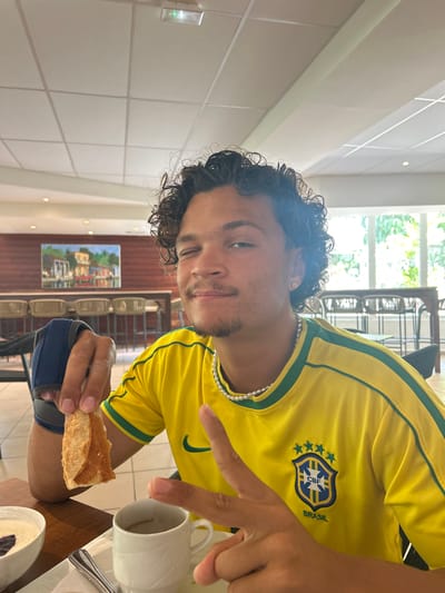 A man wearing a yellow Brazil soccer jersey and a pearl necklace is eating at a table in Sainte-Anne, France. He holds a piece of bread in his left hand and gestures with his right hand toward a white coffee cup. A painting hangs on the wall behind him.