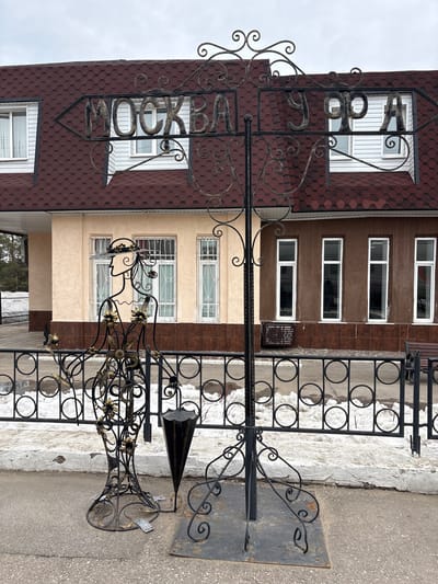 The scene in Dimitrovgrad, Russia, displays an outdoor setting featuring a dark wrought iron signpost and an adjacent, stylized wrought iron sculpture. The signpost, anchored to the asphalt pavement, includes an arrow pointing left with the Cyrillic word "МОСКВА" (Moscow) crafted in metal letters, and the Cyrillic word "УФА" (Ufa) is also present to the right, all adorned with decorative scrollwork. Next to the signpost, a wrought iron sculpture depicts a female profile wearing a hat, with the figure's body adorned with gold-colored floral accents. An inverted, dark cone-shaped object stands directly beside the sculpture.
In the mid-ground, a black wrought iron fence featuring circular patterns separates the asphalt pavement from a raised strip of ground. This strip contains patches of melting snow and degraded white concrete or plaster.
The background features a two-story building with a dark red hexagonal shingle roof. The upper level facade consists of white horizontal siding with white-framed windows. The ground level's facade is segmented: the left portion is a lighter beige stucco with white-barred windows, while the right portion is a darker brown textured wall with white-framed, tall, narrow windows. A rectangular metal ventilation unit is affixed to the lower portion of the brown wall. A dark brown wooden bench is partially visible on the far right edge of the frame. The sky above is overcast. No persons are visible within the frame.