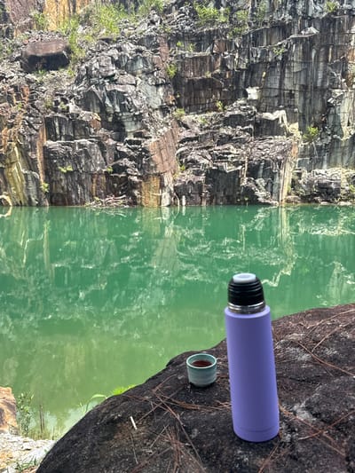 An environmental scene captures a serene moment with a purple thermos and a small ceramic cup filled with a dark liquid resting on a large, dark rock in the foreground. Beyond the rock, a vast body of calm, emerald-green water reflects the imposing, rugged cliff face of a quarry. The quarry walls are characterized by distinct, layered rock formations with variations in color, ranging from dark gray and brown to patches of ochre and white. Sparse green vegetation clings to the rocky surfaces and lines the water's edge. The presence of pine needles scattered on the rock suggests a natural, possibly wooded, environment. The location is identified as a quarry in the Lang Biang area of Da Lat, Vietnam, a region known for its natural beauty and mountainous terrain. The overall impression is one of tranquility and natural splendor, with the man-made object of the thermos and cup juxtaposed against the raw, geological landscape.