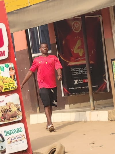 The scene is located outdoors in Puerto López, Colombia, on a light-colored ground surface. A male individual, appearing to be a young adult, walks from left to right across the frame. He wears a red short-sleeved t-shirt with a small Nike logo on the left chest, black shorts with a white drawstring and a white graphic on the left leg, and light-colored flip-flops. A necklace with a yellow circular pendant is visible around his neck. His gaze is directed forward. To the left, a red-framed vertical sign displays multiple white panels advertising food items, including "Fried Rice," "Delicious" offerings, and "Icream Supreme." The background features a building with a reddish-brown and dark brown facade. A large red banner on the building advertises a "4 ANNIVERSARY," with text thanking for "support and patronage" and referencing "30 November 202[X]." A dark window is visible on the building. Above the building, a bright yellow overhang or structure is present. A light-colored, discarded tire rests on the ground in the lower right foreground.