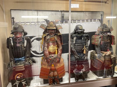 This image displays a museum exhibit in Iga, Japan, featuring four samurai armors, each set in a glass display case. The armors are positioned from left to right, with the first one having a red face mask and a dark helmet with a stylized crest. The second armor is a vibrant red and gold ensemble with a prominent chest plate and elaborate leg coverings. The third armor is primarily black, with a dark helmet and intricate detailing on the chest and arms. The fourth armor, partially visible on the far right, also appears to be dark with red and gold accents. The background of the exhibit consists of a large screen with a black and white circular motif and horizontal bands of blue and white patterns, suggesting a historical or cultural theme. Various labels and descriptive texts are placed around the display, providing information about the artifacts. The lighting within the exhibit is bright, illuminating the armors and their intricate craftsmanship.
