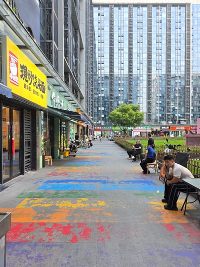 This image depicts a street scene in Linping District, China. The setting is an urban environment with a modern, glass-fronted building on the left, likely an office or commercial complex. In the foreground, a paved walkway is decorated with colorful, painted stripes resembling a rainbow. Several people are present, some seated at tables and chairs placed along the walkway, suggesting an outdoor dining or social area.

On the right side of the walkway, a few individuals are interacting. One person, wearing a white t-shirt and black pants, is leaning forward, possibly engaged in a conversation or activity at a table. Another person in a blue shirt is also seated. Further down the walkway, more people are visible, some appearing to be seated as well.

Several shops or establishments are visible along the right side of the walkway, indicated by signs and storefronts. One prominent yellow sign with Chinese characters stands out, featuring a red graphic of what appears to be a bowl of noodles. Other green and darker colored signs are also present.

The time of day appears to be daytime, with bright lighting casting some shadows. The weather seems pleasant. The overall atmosphere appears relaxed and casual, with people enjoying the outdoor space. The rainbow-colored pavement adds a vibrant and cheerful element to the scene.