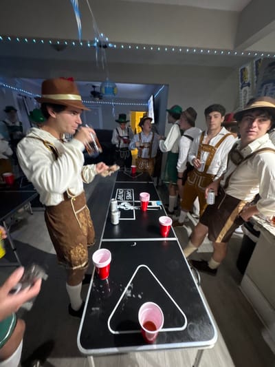 This image depicts an indoor social gathering of approximately ten young adult males in a residential setting in South Bend, United States. All identifiable individuals are dressed in Bavarian-themed attire, consisting of lederhosen-style shorts or pants, white or cream-colored long-sleeved shirts, and various hats including brown fedoras and green alpine hats.
The central foreground features a male subject in a brown fedora and brown lederhosen, consuming liquid from a dark bottle and holding a small, flat food item. Immediately to his right, a black folding table, set up for beer pong, displays white markings for cup arrangement, including two triangular outlines. Multiple red plastic cups, some containing liquid, and two silver beverage cans are present on this table.
On the right side, another male subject in similar attire, wearing a brown hat, stands with a silver beverage can, looking directly toward the camera. Additional males in themed costumes are positioned in the mid-ground and background, standing and engaged in conversation or observation.
The environment includes blue and white decorative streamers and string lights suspended from the ceiling. A blue and white patterned banner is affixed to a wall on the right. A dark sofa is partially visible in the distant background. The flooring is a light-toned hard surface. The scene portrays a themed party atmosphere.
