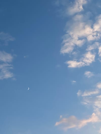 This is a vertical, eye-level shot of a clear blue sky with scattered white and pinkish clouds. A crescent moon is visible in the upper left quadrant of the sky. There are no people or objects other than the celestial bodies and clouds in the image. The environment is an outdoor sky scene. No specific location in Ilavche, Ukraine is identifiable from the image.