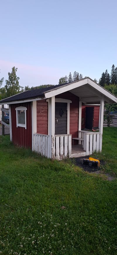 The image captures a small, weathered red wooden cabin with a dark-colored door adorned with a diamond-shaped detail and a silver handle. The cabin features a white-trimmed window on its left side. A white, peeling wooden fence encloses a small porch area leading to the cabin's entrance, which includes a wooden step and a built-in bench. To the right of the porch, another dark-colored door is visible. The cabin sits on a patch of green grass, with a small yellow toy truck overturned on a black mat in front of the porch. In the background, trees line a hill, and the sky is a pale blue, suggesting it is either early morning or late evening. The overall setting appears to be a rural or suburban environment, possibly a backyard or a small park area within or near the city of Salpakangas, Finland, given the presence of coniferous trees and deciduous trees typical of the region.