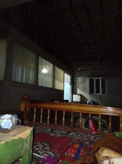 This is an indoor, low-light photograph taken at night, likely in a residential setting. The perspective is from a seated or lying down position, looking across a wooden railing with decorative spindles. To the left, on a green-colored surface, rests a translucent glass mug with a blue and white floral design. The railing itself is made of polished wood and extends across the frame.
Beyond the railing, the scene depicts a somewhat cluttered room. A red rug with a black and white floral pattern covers the floor. In the background, there is a white appliance that resembles a washing machine or a chest freezer, situated near a wall. To the right of this appliance, a white window with multiple panes is visible, reflecting light.
Further into the background, a doorway or opening leads to another space, where additional items and possibly another appliance are partially visible. The ceiling is constructed with exposed wooden beams and metal framing, suggesting an unfinished or utilitarian structure. The overall impression is of a lived-in, possibly storage or multipurpose area.
The specific location in Khamza, Uzbekistan, cannot be determined from the visual information alone.