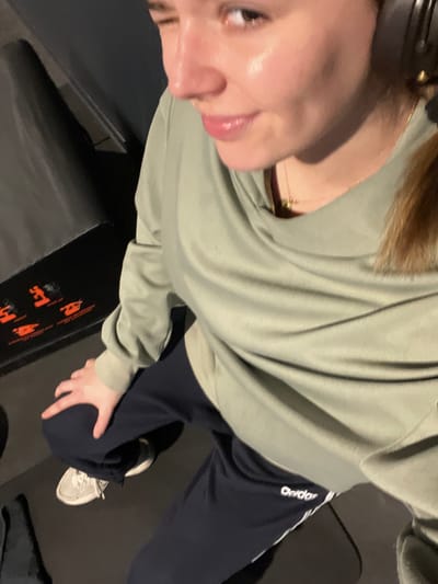 An image captures a female individual engaged in physical activity within an indoor setting in Annœullin, France. The subject, viewed from a slightly elevated perspective, has a fair complexion, brown hair, and wears black over-ear headphones, a light green long-sleeved top, dark blue Adidas sweatpants with white side stripes, and white athletic footwear. Her face exhibits a sheen consistent with perspiration, her left eye is closed in a wink, and her lips are slightly parted in a faint smile. A thin gold-colored necklace is visible. She adopts a crouched posture, with her left hand resting on her left knee. In the foreground to her left, a dark-colored piece of exercise equipment with orange graphic indicators is visible on a dark floor. The background is indistinct and dark.