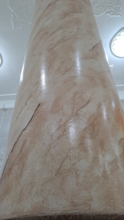 The image shows a close-up of a textured column with a marble-like finish. The column is curved and appears to be part of a larger structure, possibly an interior space. The texture is characterized by swirling patterns of light brown and beige, with some darker brown accents that resemble natural stone veins. The surface is glossy, reflecting light in irregular patches.
In the upper left and lower left of the image, there are glimpses of white architectural elements, including what appear to be decorative moldings and an oval light fixture. These elements suggest an indoor setting with some form of ornate design. The lighting appears to be coming from the fixtures, creating highlights and shadows on the column's surface, indicating it might be daytime or a well-lit interior.
The texture on the column shows a variety of brush strokes or application techniques, creating a dynamic and uneven surface. There are areas with fine, linear textures and others with a smoother, blended appearance. A thin, dark crack is visible on the surface of the column, running diagonally across a section of the image. The right edge of the image shows a darker, rougher texture, possibly a carpet or a different material adjacent to the column.
There is no visible text in the image. The focus is entirely on the architectural detail and the tactile quality of the column's finish. The overall impression is one of a detailed study of an interior architectural element.