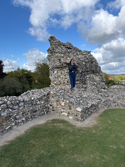 An outdoor, eye-level shot depicts a person lying on a rugged, ancient stone wall under a partially cloudy sky. The image is oriented vertically, with the ground plane to the right and the sky filling the upper left.
The main subject, a person, is dressed in a black leather jacket, dark blue jeans, and dark shoes, with blue socks peeking out. They are wearing sunglasses and have dark hair. They are positioned on the stone wall, appearing to lean or lie back, with their left arm bent and supporting them against the wall and their right arm extended outwards. They are looking towards the camera with a relaxed expression.
The stone wall, likely part of a ruin or fortification, is prominently featured, constructed from numerous irregular grey and dark-colored flint stones set in mortar. It forms a textured, uneven surface on which the person rests. To the right of the wall, a strip of worn path transitions into a lush green grassy area, suggesting a well-maintained outdoor space.
In the background, beyond the wall, are green trees and foliage, with glimpses of distant buildings and structures, hinting at a village or town. The sky is a vibrant blue, adorned with large, fluffy white and grey clouds, indicating a bright, possibly partly cloudy day. The overall atmosphere is pleasant and casual, with the scene well-lit by natural daylight. There is no visible text in the image.