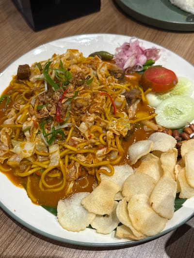 A close-up overhead shot shows a white plate of noodles with various accompaniments on a wooden table. The main dish is a mound of yellow noodles coated in a rich, orange sauce, mixed with shredded cabbage and topped with fried shallots and green onions. To the right of the noodles are slices of cucumber, a wedge of tomato, and a small pile of pickled red onions with a few green chilies. In the lower right corner, a scattering of peanuts and several light-colored crackers are visible. The plate has a subtle green rim. In the background, out of focus, a dark rectangular object is partially visible on the left, and a green plate with a fork is on the right. The lighting is dim, suggesting an indoor setting. There are no people or animals in the image. The scene is likely a restaurant in Gunung Putri, Indonesia, given the provided context.