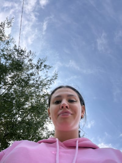 An outdoor, low-angle, selfie-style shot shows a young adult with dark hair and makeup, wearing a pink hoodie. The subject's face is partially visible from the chin up, with their head tilted back and looking towards the camera with a slight smile. Their eyes are looking up, and they have dramatic eye makeup, including false eyelashes. The background consists of a bright blue sky with scattered white clouds and the upper branches of a leafy green tree on the left side of the frame. A thin dark wire runs vertically through the upper left portion of the image. The location is described as Chumna, Russia.
