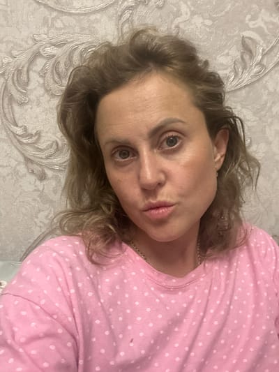 A woman with blonde hair and brown eyes is shown in a close-up portrait. She is wearing a pink polka dot shirt and has a chain necklace on. The background is a textured wall with a decorative pattern. The image appears to be taken indoors, possibly in a residential setting in Verkhniy Yurt, Russia.