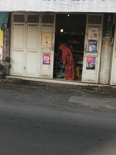 A person wearing an orange hijab and a patterned orange dress is sweeping the ground in front of a store entrance in Glagah Arum, Indonesia. The store has a white double door with vertical slats at the top. Advertisements are posted on the doors and the wall. The ground in front of the store is paved with asphalt and there are bricks lined up near the entrance.