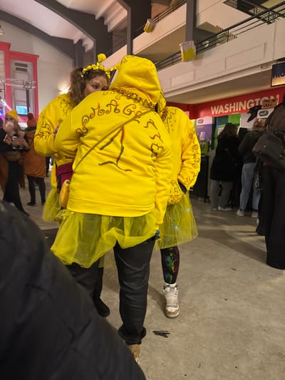 Two people are dressed in bright yellow hooded sweatshirts with gold necklaces and yellow tulle skirts. One person has a floral headpiece. They are in a crowded indoor setting with concrete floors. Behind them is a red banner with "WASHINGTON" written on it. Other people are visible in the background, some wearing jackets and jeans. The lighting appears to be artificial and somewhat dim.