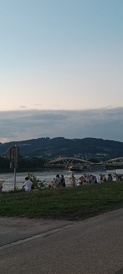 The image depicts a serene evening scene along the Danube River in Linz, Austria. The sky is a gradient of pale blue to a softer, lighter hue near the horizon, with subtle cloud formations adding texture. In the mid-ground, a dark, forested hill rises, dotted with faint lights of a settlement, and a distinct yellow-lit bridge spans the river, with a cruise ship passing beneath it. The foreground is a grassy embankment sloping down to the river, where a group of people are seated and lounging in chairs, enjoying the view. A paved path runs along the edge of the embankment. The overall impression is one of peaceful recreation and scenic beauty.