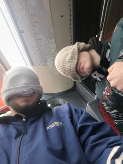 Two individuals wearing beanies pulled over their faces are visible on what appears to be public transportation. One person is wearing a blue jacket. A route map is visible on the wall of the vehicle, with sunlight streaming in. The other person is wearing a black hoodie with white lettering and a patterned jacket. The perspective suggests the photo was taken by one of the individuals.