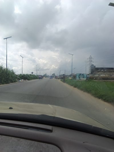 The image depicts a road scene in Yenegoa, Nigeria, under a cloudy sky. The road appears to be a multi-lane highway, with traffic visible in the distance. On the left side of the road, there is a dense growth of tall green vegetation, with some scattered refuse. Several modern streetlights with solar panels are installed along the roadside, suggesting this is a developed area.
Further down the road, there are several vehicles, including what appears to be a small bus or van and some smaller cars. A few people are visible walking along the side of the road or near parked vehicles. On the right side of the road, there are more structures, including what looks like a factory or industrial building with a tall transmission tower and power lines nearby. A blue sign with some white text is also visible on this side. The overall impression is of a busy but somewhat unkempt urban environment. The lighting suggests it could be daytime, but the heavy cloud cover gives the scene a somewhat subdued atmosphere. The presence of the transmission tower and the industrial buildings hint at an area with significant infrastructure.