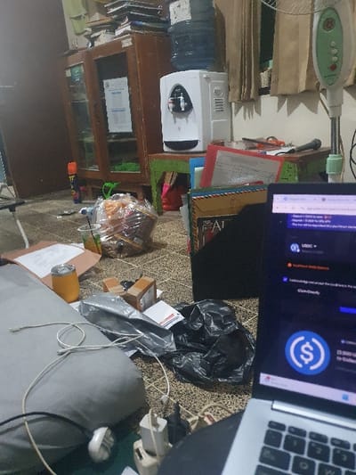 The image shows a cluttered room in Kecamatan Jombang, Indonesia, located at latitude: -7.519, longitude: 112.236. There is a wooden cabinet with glass doors, which is filled with various items. Next to the cabinet, there is a white water dispenser, a small table with a plastic bag of food, and a black laptop computer open on the floor. The laptop screen displays a website with a blue circle containing a dollar sign. A gray pillow lies on the floor, next to a small orange container. Several cords and other miscellaneous items are scattered around the room.