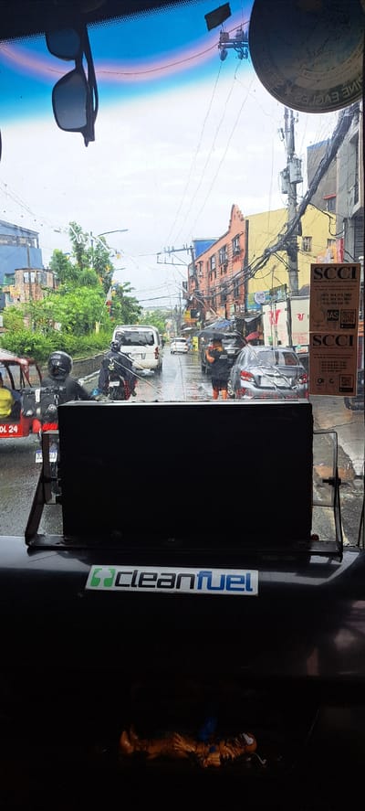 The image captures a view from inside a vehicle looking out onto a street in Mandaluyong City, Philippines. It appears to be a rainy day, with wet roads and some vehicles using umbrellas. In the foreground, a black rectangular object is mounted, possibly a screen or dashboard component, with a "cleanfuel" sticker attached to its lower edge.

Further out, the street is moderately busy with traffic. Several vehicles are visible, including a white van, a silver sedan, and a motorcycle with two riders wearing helmets and raincoats. Pedestrians are also present, some holding umbrellas. The buildings lining the street are a mix of old and new, with balconies and electrical wires forming a complex network overhead. Trees and greenery are visible on the left side of the street. Two stickers, both with "SCCI" branding, are affixed to the right side of the vehicle's window. The overall atmosphere suggests a typical urban scene during a rain shower.