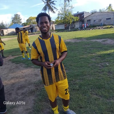 A young man in a black and yellow striped soccer jersey is standing in the center of a grassy field, holding a small round object in his hands. The field is located in Nungwi, Tanzania (latitude: -5.726, longitude: 39.301). There are other soccer players wearing the same jersey in the background. In the distance, a low building with a tiled roof and a small palm tree can be seen. The sun is shining and the sky is clear. The grass is green and there are some patches of brown dirt. The young man is smiling. The image was taken with a Galaxy A05s phone.