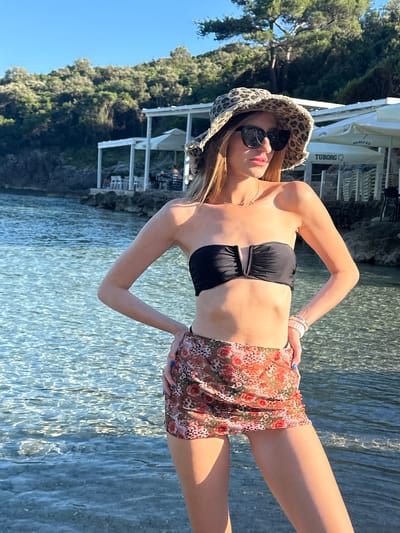 A young adult female stands in clear, shallow water, likely a cove or beach, in Kubasi, Montenegro. She is positioned with hands on hips, body slightly angled right, and head turned left. She wears a black strapless bandeau bikini top, a sheer floral and leopard print mini-skirt, a leopard print bucket hat with a frayed brim, and dark sunglasses. A delicate gold-toned necklace and multiple light-colored bracelets on her right wrist are visible, along with blue nail polish on her fingers. The water's surface is rippled, revealing a rocky bottom. In the midground, white open-air structures with rectangular pillars, tables, and chairs line a rocky shore. A "TUBORG" sign is visible on a white umbrella or awning among these structures. Dense green Mediterranean vegetation covers the hills behind the structures. The sky is clear blue, indicating strong sunlight.