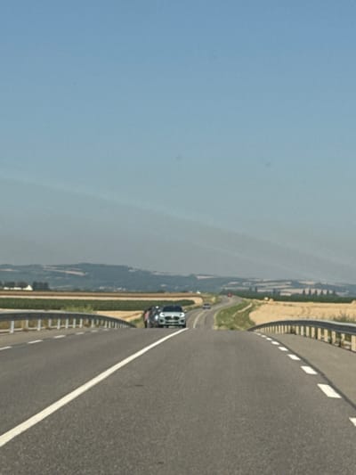 A curved highway dominates the frame, a white line marking its edge. A vehicle drives along the road, and another smaller road with a car visible in the distance runs parallel. Beyond the road, a green landscape transitions into a hazy sky. The road surface is asphalt, and a guardrail borders the highway's edge. The image has been rotated.