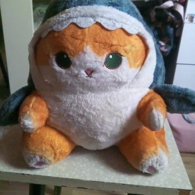 A plush toy, resembling a cat wearing a shark costume, is positioned centrally on a white, patterned table. The toy is predominantly orange and white, with blue accents from the shark hood. Its eyes are large and green, and it has a small pink nose. The background is blurred, showing hints of furniture and possibly a window. The image appears to be taken indoors, likely in a residential setting. The location of Tomilino, Russia is not discernible from the visual information present in the image.