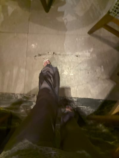 A first-person perspective shows a person standing on a tiled floor. One foot is visible, clad in a gold sandal and dark fabric covering the leg. Part of a wooden chair leg is in the upper left corner. The background consists of more floor tiles, and some green shrubbery is visible along the right side. The lighting appears to be dim, possibly evening.