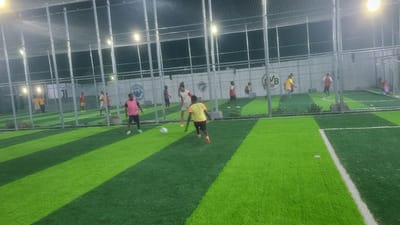The image depicts an outdoor, artificially lit futsal or small-sided football match taking place at night on an artificial turf pitch in Babawa, Nigeria. The pitch features alternating strips of light green and dark green turf, marked with white boundary lines. The entire playing area is enclosed by a tall metal fence with chain-link netting. Multiple overhead lights illuminate the scene, casting distinct shadows and bright reflections on the playing surface.
Approximately 10-12 male individuals, appearing to be young adults, are present on the pitch, engaged in or observing the game. In the foreground, slightly to the right of the center, a player wearing a bright yellow/lime green t-shirt and black shorts is actively kicking a white soccer ball towards the right side of the frame. He has orange athletic footwear. Adjacent and slightly behind him, another player in a white t-shirt and light shorts observes the ball. To the far left, a player in a pink vest and dark shorts stands with white athletic shoes. Other players are scattered across the pitch, dressed in various colored athletic wear including red, yellow, and blue shirts, with one individual near the right goal appearing to be a goalkeeper.
A light-colored wall or building forms the background behind the fence. Several circular logos are visible on this background wall through the fence: a blue "Manchester City FC" crest is visible on the left, while a darker logo featuring the letters "VB" and numbers "09" is seen towards the center-right. Another smaller, darker circular emblem with a crown-like symbol is present near the center. A white goal frame is visible on the far right of the pitch. Minimal vegetation is present at the base of the fence posts.