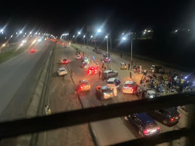This media file captures a nighttime scene, likely in Sauka, Nigeria, that appears to be a car accident or a major disruption on a road. The image is taken from an elevated perspective, looking down on a highway or a large road.
The primary subjects are numerous vehicles, many with their brake lights illuminated, suggesting they have stopped abruptly. Several cars are visible, some appearing to be involved in a collision or stopped due to an obstruction. In the foreground and middle ground, there are groups of people gathered on the road, some appearing to be onlookers or emergency responders. They are illuminated by the lights of the vehicles and streetlights.
The scene is set at night, as indicated by the darkness and the prominent vehicle lights. The presence of streetlights and the general road infrastructure suggest an urban or semi-urban environment. The activity taking place seems to be a chaotic halt of traffic, with people congregating on the road. It's difficult to ascertain specific weather conditions, but the image appears clear. There are no discernible emotions that can be clearly identified due to the distance and low light.
Notable details include the presence of what appear to be utility poles with lights, illuminating sections of the road. The sheer number of people and vehicles suggests a significant event. The image is somewhat blurry, possibly due to low light conditions, movement, or the quality of the recording device. There is no visible text in the image.