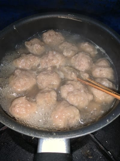The image shows a close-up, overhead view of meatballs being boiled in a pot. The pot is dark-colored with a black handle. The meatballs are pale brown and irregular in shape, with some appearing to be slightly larger than others. They are submerged in clear, bubbling water, indicating they are actively cooking. A pair of wooden chopsticks is visible in the center of the pot, resting on top of the meatballs and submerged in the water, suggesting they are being used to stir or check the cooking process. The surrounding environment appears to be a kitchen, with a dark, textured surface beneath the pot, possibly a stovetop. The lighting is somewhat dim, but enough to clearly see the details of the food and the pot. The overall impression is that of a home-cooked meal being prepared. There is no visible text in the image. The location context of Karangkemiri, Indonesia, suggests this is likely a common culinary practice in that region.