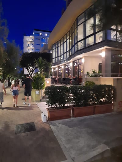 The image captures a scene in the city of Pesaro, Italy, likely during the evening. In the foreground, a paved walkway is visible, with a drain cover set into the bricks. To the right, two white plastic containers are placed on the walkway. Further along the walkway, two individuals, a man and a woman, are walking away from the camera. The man is wearing a black t-shirt and shorts, while the woman is wearing a pink tank top and denim shorts. The walkway leads to a street lined with trees and a building with large windows, suggesting it could be a commercial area or restaurant. Behind the trees and walkway, a modern multi-story building is illuminated, with its windows glowing against the dark blue night sky. The overall atmosphere is that of a relaxed evening stroll in an urban setting.