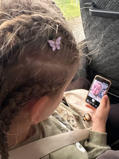 The image displays a close-up, over-the-shoulder view of a child seated within a vehicle, observed from the child's right side. The provided image does not contain visual information to confirm the specified location in the city of Кварса, Russia.

The child has light brown hair styled in braids, with numerous fine, loose strands. A small, light purple butterfly-shaped hair clip is affixed to the hair on the right side of the child's head. The child's right ear is visible. The child is wearing a light green long-sleeved top featuring a subtle pattern, possibly of animal faces or similar motifs. A light-colored seatbelt diagonally crosses the child's torso.

The child's right hand holds a smartphone with a metallic gold or bronze frame. The phone's screen displays a front-facing camera view, showing the reflection of a person, possibly an adult, with light-colored hair, making a playful facial expression with their tongue extended. On the child's lap or clothing, below the phone, a small Funko Pop figurine with brown hair and a striped top is visible.

The background indicates an interior setting, likely a vehicle such as a car or bus. To the left, a window shows blurred green foliage and bright daylight. To the right, a dark, textured interior panel of the vehicle with a visible dark handle is present. The seating material behind the child appears to be a dark, textured fabric.