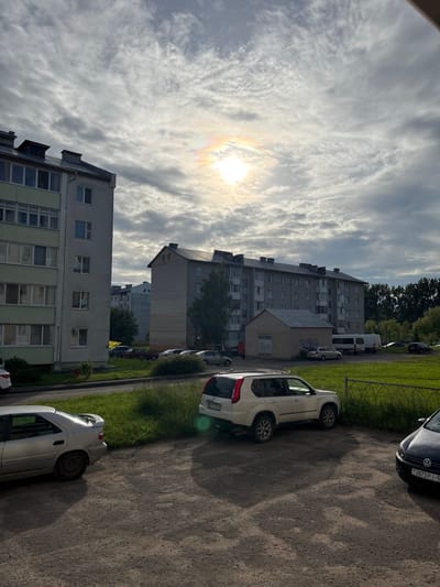 The image captures a residential scene in Vileyka, Belarus, likely taken in the late afternoon or early evening due to the low angle of the sun and long shadows. The weather appears partly cloudy with dramatic, textured clouds across a bright sky. The sun itself is visible, partially obscured by clouds, creating a faint rainbow effect.
The foreground features a gravel road or parking area with several cars parked along it. A white SUV is prominently parked on the right side of the image. Further down the road, a silver car is visible, and another white vehicle, possibly a van, is parked in front of a brick building. A dark-colored car with a visible license plate is parked on the left side, with some text on its rear.
Behind the parked cars, there is a strip of green grass and a low metal fence. Several multi-story residential buildings dominate the background. The closest building on the left is a light-colored structure with many windows, some of which appear to have a greenish tint. To its right, a taller, grey brick building with numerous windows and visible roof structures stands. Further in the distance, more buildings and trees are visible, suggesting a typical urban or suburban residential area. The scene conveys a sense of everyday life in a Belarusian town.
