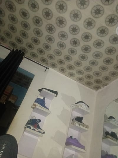 The image depicts an indoor scene in Ogbogbo, Nigeria, characterized by a distinct ceiling pattern and a display of footwear.
The ceiling is covered with a light-colored material, possibly plaster or tile, featuring a repetitive grid pattern of dark gray circular motifs. Each circle contains an eight-pointed star design. The pattern is consistent across the entire visible ceiling surface, extending into the corner where it meets a plain white wall.
The primary wall on the right is painted white and supports two vertical shelving units. Each unit consists of multiple rectangular white shelves.
On the left shelving unit, a blue high-top boot with a tan/white sole is placed on the top shelf. Below it, a dark blue, white, and possibly red high-top sneaker rests on the second shelf.
The right shelving unit displays several items. The top shelf contains a stack of white material, potentially clothing, topped by a dark fabric item. The second shelf holds a beige or off-white high-top sneaker with dark laces. Below this, a purple low-top sneaker is situated on the third shelf. The lowest visible shelf on this unit is partially obscured.
To the far right, a tall, arched mirror reflects portions of the shelving units and the shoes displayed on them.
On the left side of the image, a black curtain is drawn, covering a doorway or window opening. A black curtain rod extends horizontally above it. Through the opening, a light blue wall is visible in the background, with indistinct rectangular objects (possibly boxes or furniture) appearing in muted tones.
In the lower-left corner, the left shoulder and upper arm of a person are partially visible. The person is wearing a dark gray garment with the letters "ORA" visible, suggesting part of a brand name. The rest of the person is outside the frame. The overall lighting is dim, suggesting an indoor setting with either limited natural light or artificial illumination.