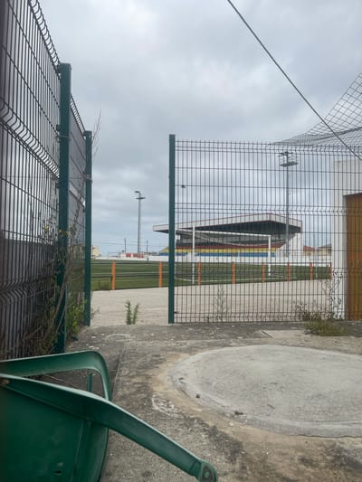 An exterior, eye-level, medium shot captures a view through a fence towards a sports stadium under a cloudy sky. In the foreground, a green metal chair is partially visible, tipped over on a concrete surface. The ground is a mix of concrete and gravel, with small weeds growing through. A dark metal fence with green posts runs along the left side of the frame, and another fence with green posts and wire mesh forms an opening in the center, leading to the stadium. The stadium features a grandstand with multi-colored seating in red, yellow, and blue, and a white roof structure. Behind the stadium, tall light poles and a few buildings are visible. The overall scene suggests an abandoned or disused sports facility. The location is identified as São João das Lampas, Portugal.
