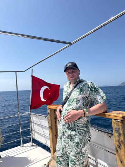A medium-angle shot shows a man wearing a leafy green and white Hawaiian-style shirt, a black Nike baseball cap, and a black shoulder strap, on a boat with the Turkish flag waving behind him on a clear, sunny day. The man is leaning against a wooden ledge, with his left hand resting on his stomach. He is looking directly at the camera. He has two wristbands on his left wrist, one green and one orange. The boat is moving on a body of water, likely the sea, and there is a clear blue sky in the background. The boat has a metal railing system and a partially visible white awning. The city is Unknown, Türkiye.