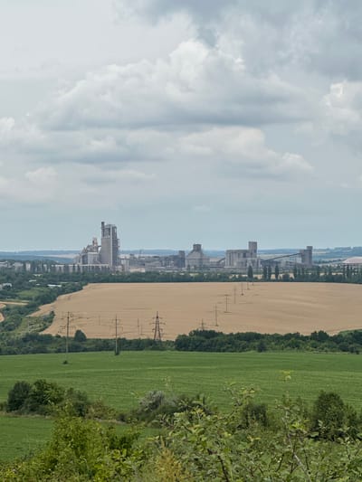 This is a wide shot of an industrial facility located in Kolubaivtsi, Ukraine. The facility, which appears to be a cement plant, is situated in the background, characterized by its tall silos, conveyor belts, and various industrial structures. In the mid-ground, there is a vast field of dry, light-brown crops, possibly wheat or barley, with a line of tall utility poles running through it. The foreground features a lush, green field, suggesting agricultural land. The sky is overcast with a mix of light and dark clouds. The overall scene depicts a rural landscape with a prominent industrial presence. There are no people or animals visible in the image.