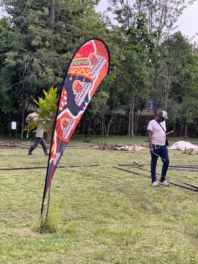 The image captures an outdoor scene, likely in a field or park, characterized by a vast expanse of green grass and numerous trees under an overcast sky. The setting appears to be Makuyu, Kenya.
In the left foreground, a prominent, colorful teardrop-shaped banner stands upright. The banner's design is vibrant, featuring traditional-looking patterns in shades of red, orange, yellow, and black on a dark background. Repeated text on the banner clearly reads "GUCHOKIA RUI MUKARO," with a smaller line below specifying "A Reggae Culture Festival." Graphic elements resembling stylized calabashes or drums are also visible on the banner.
Towards the center-right of the frame, a man stands facing to his left. He wears a light-colored t-shirt, dark trousers (possibly jeans), white sneakers, and a light-colored baseball cap. A shoulder bag is visible across his body. Further back and partially obscured by the banner and some foliage, another individual can be seen, possibly tending to a small plant.
The grassy ground extends into the background, where numerous wooden poles or beams are scattered, suggesting the setup or dismantling of structures for an event. The backdrop is filled with lush green trees, indicating a natural environment. The overall impression is that of an outdoor event, specifically a reggae culture festival, in its preparatory stages during the daytime.