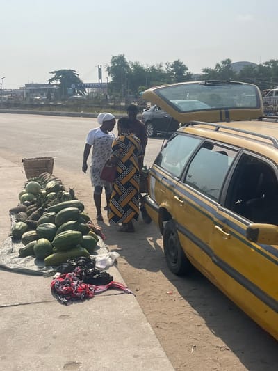 The scene depicts a roadside market setting in Ikota, Lagos, Nigeria, under bright daylight conditions. A blue sign reading "VICTORIA ISLAND" is visible in the background. On the left, a large display of fruits, including numerous green papayas, pineapples, and spherical green fruits (possibly watermelons or gourds), rests on a grey tarp on the ground, adjacent to a concrete curb. A woven basket is positioned near the top of the fruit pile. Below the fruit display, black plastic bags and crumpled red-and-white patterned fabric are situated on the concrete curb.
Three individuals are gathered to the right of the fruit stall. A woman wearing a white head covering, a white short-sleeved top, and a light-colored patterned wrap stands facing right, seemingly in conversation. She faces a second woman, whose back is largely to the camera, wearing a long dress with yellow, black, and white geometric patterns and carrying a small reddish-brown handbag. A man in a dark top is partially visible behind the second woman. To the far right, a yellow station wagon, featuring blue stripes along its side, is parked on a dirt shoulder with its rear hatch open, exposing the cargo area. A paved road stretches behind the individuals and the fruit display, with distant buildings, trees, and other vehicles visible under a hazy sky.