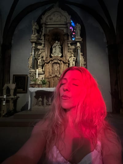 In the dimly lit interior of the Church of the Assumption of the Blessed Virgin Mary in Vianden, Luxembourg, a woman is captured in a selfie, her face illuminated by a striking red light. Her eyes are closed, and her lips are slightly parted, suggesting a moment of introspection or meditation. The vibrant red light casts a dramatic glow across her blonde hair and shoulders, creating a stark contrast with the white lace of her garment.

Behind her, the church's ornate altar stands as a testament to its historical and religious significance. Sculptures of figures, likely religious in nature, adorn the elaborate wooden structure. A stained-glass window, partially visible in the background, adds subtle touches of blue and red light to the scene. The architectural elements of the church, including arched ceilings and stone columns, are discernible in the shadows. To the left of the altar, a framed painting hangs on the wall, its details obscured by the low light. The overall atmosphere is one of solemnity and perhaps a touch of mystique, intensified by the unusual lighting and the subject's contemplative pose.